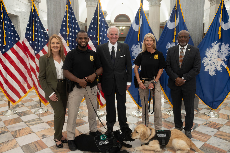 Five people dressed in formal and law enforcement attire stand indoors on a marble floor with American and South Carolina flags behind them, accompanied by two electronic detection canines wearing vests.