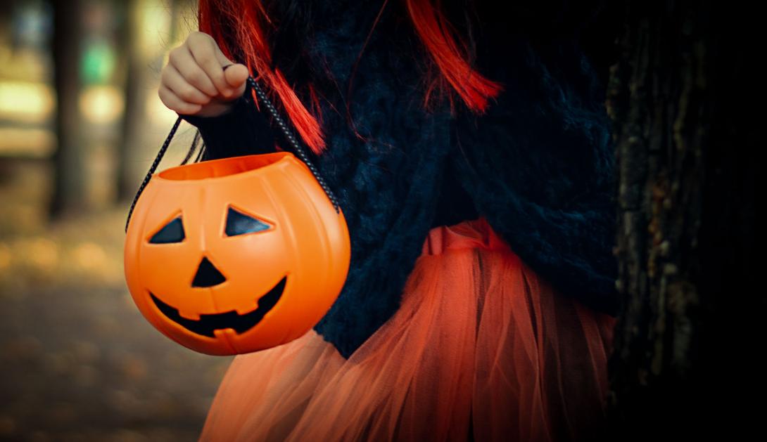 Happy girl wearing a witch's hat, with a pumpkin handbag, peeps out from behind a tree.