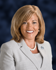 Portrait photo of a smiling white woman with blonde hair, wearing a gray suit and a white pearl necklace.